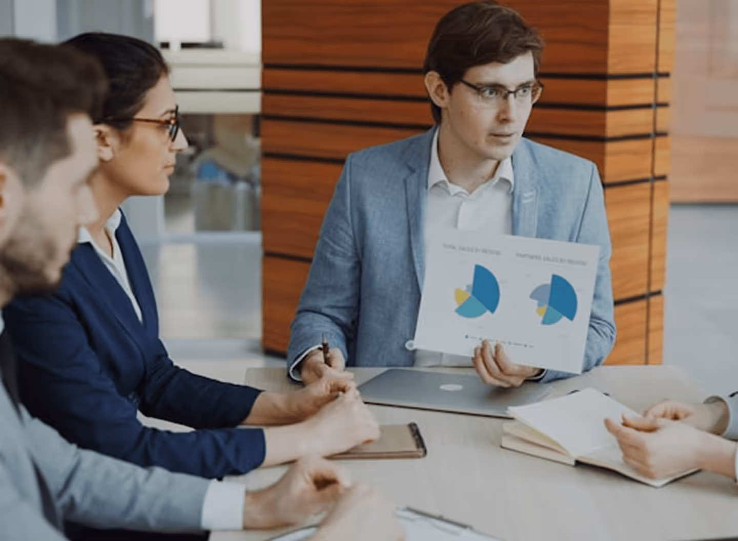 Businessman presenting pie charts to colleagues at a meeting table with notebooks and a laptop.