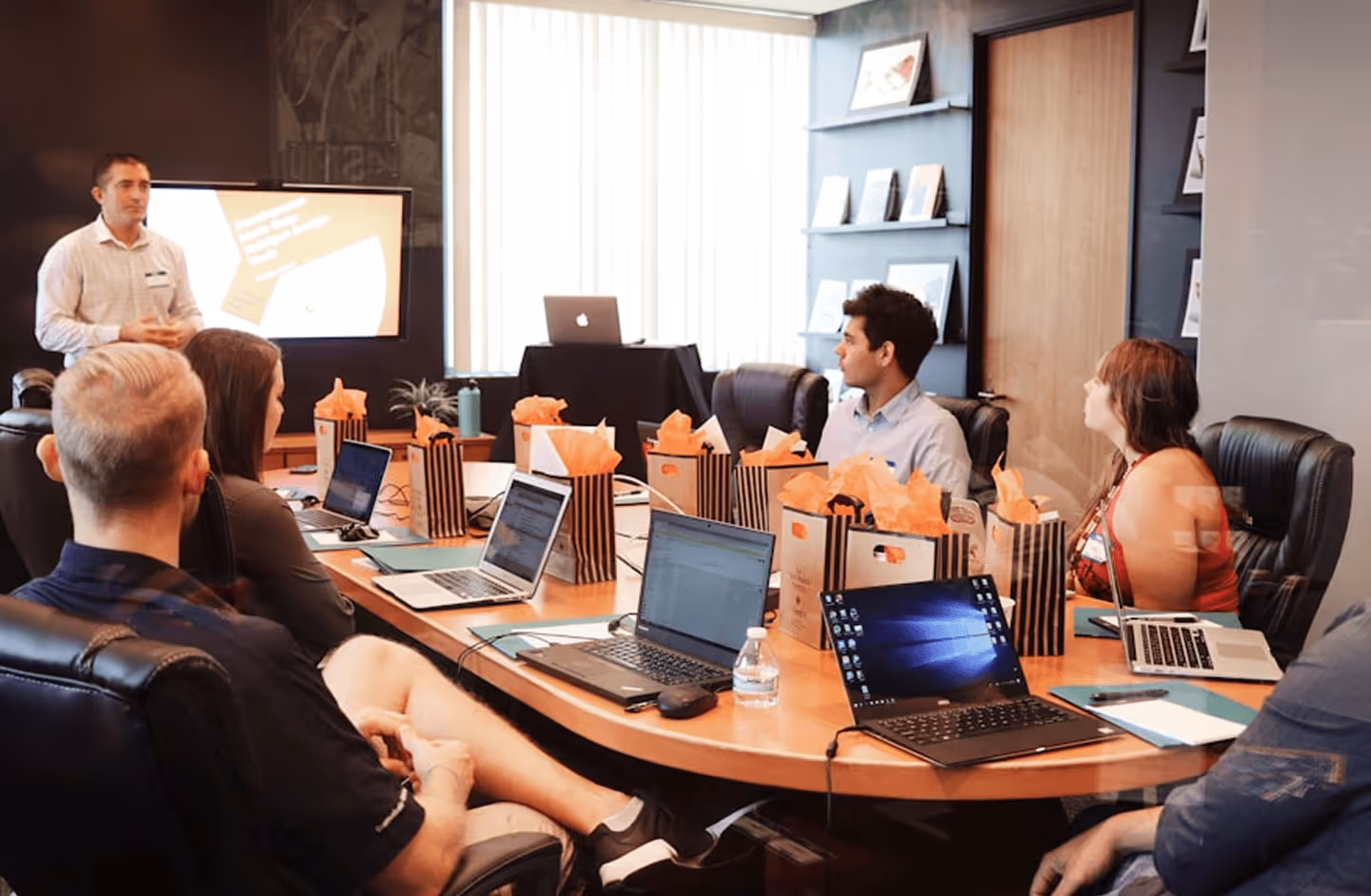 Group of six people in a meeting room with laptops and gift bags, one man presenting near screen.