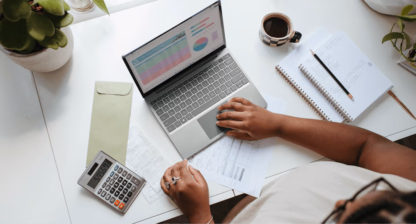 Person using laptop and calculator at desk with papers, notebooks, pencil, coffee cup, and plants nearby.