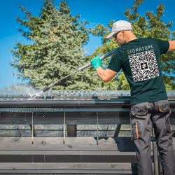 Man in cap pressure washing a metal rooftop outdoors with trees in the background.