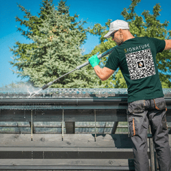 Man in cap pressure washing a metal rooftop outdoors with trees in the background.