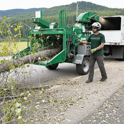 Man feeding a large tree branch into a green wood chipper on a rural road near a truck.