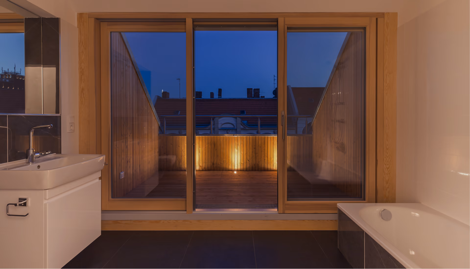 Badezimmer mit Holzfensterrahmen und Blick auf beleuchtete Dachterrasse mit Betonelementen am Abend – Projekt Dachaufstockung Berlin
