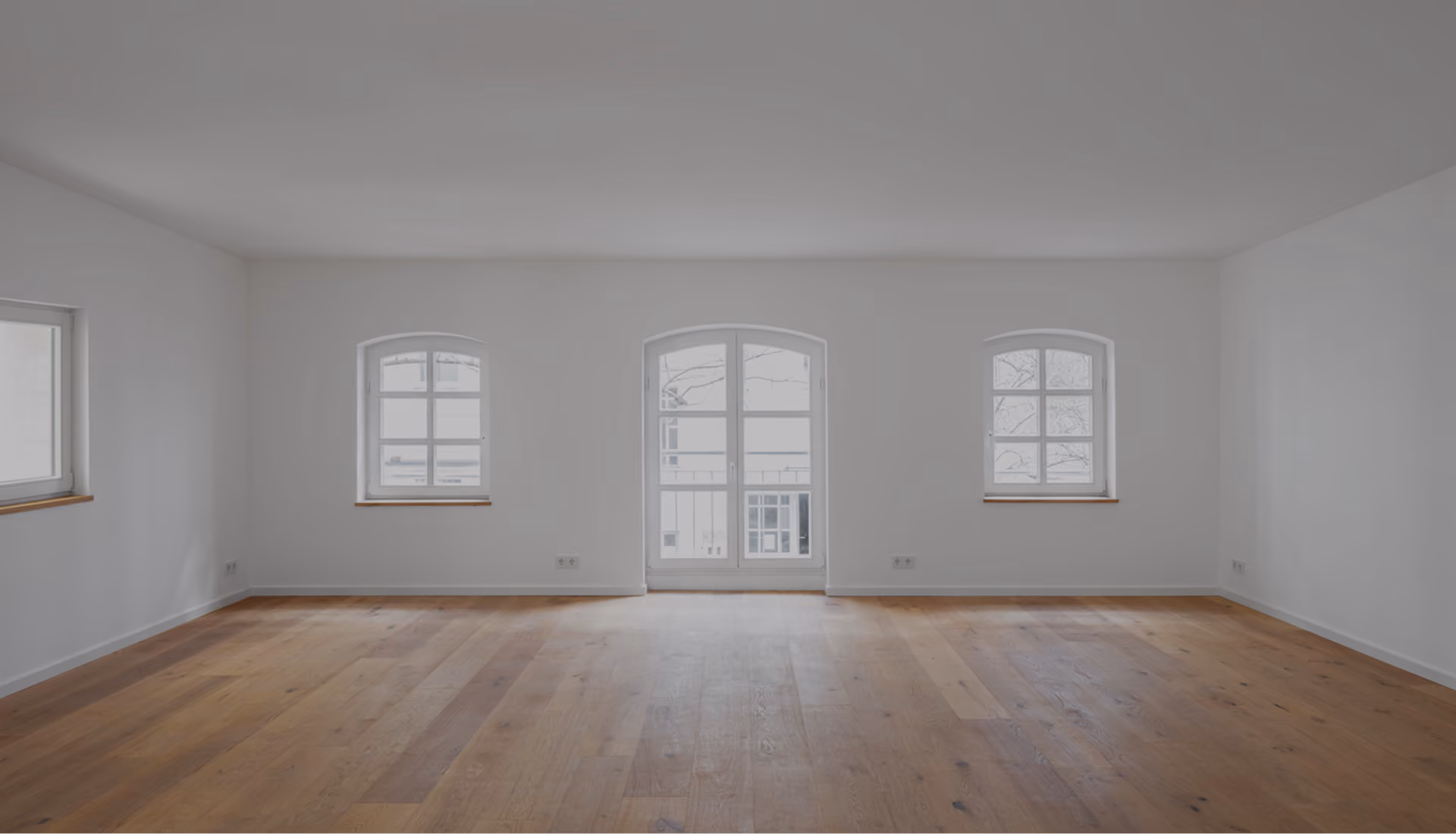 Leerer Wohnraum im Projekt Remise mit drei unterschiedlich geformten Altbaufenstern und hellem Dielenboden