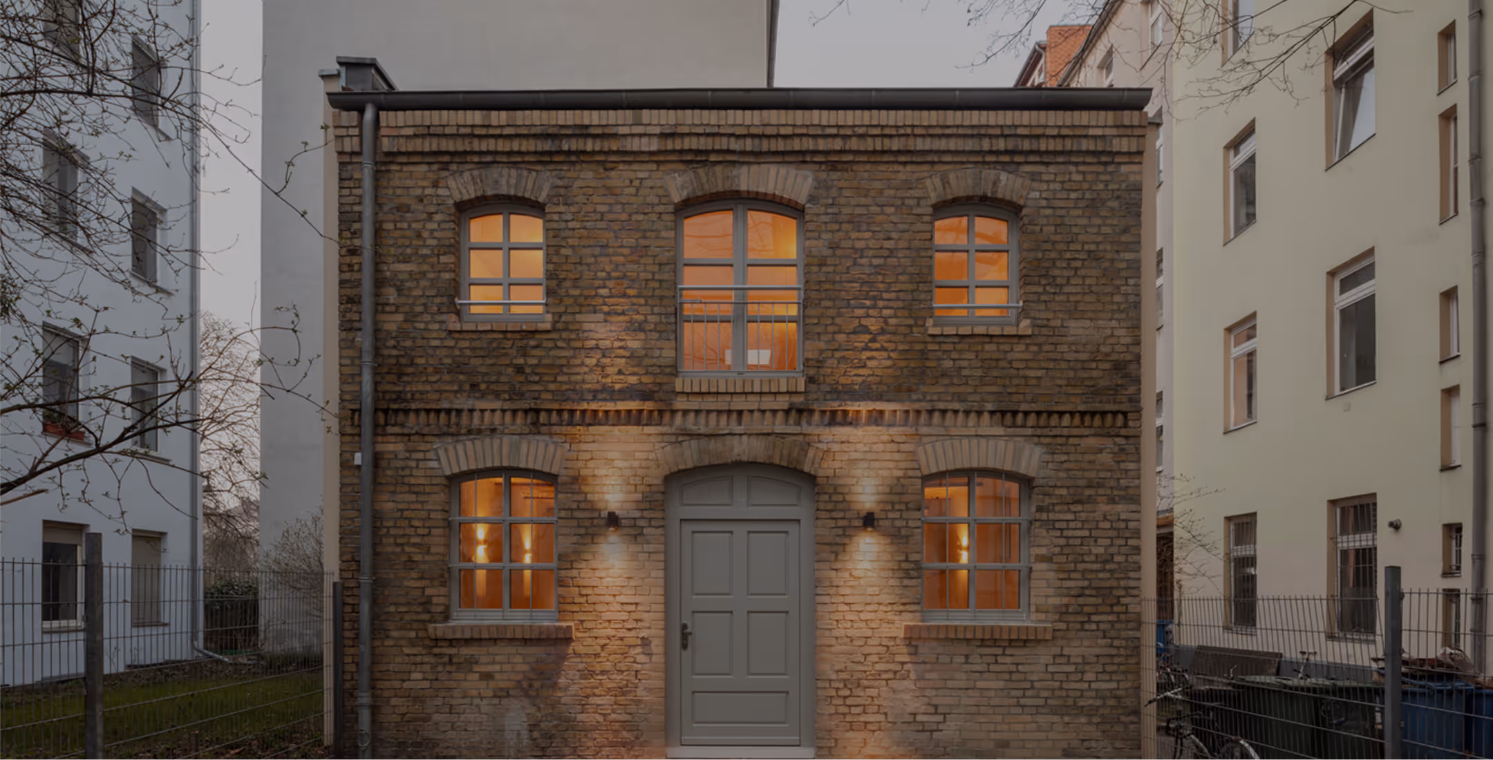 Sanierte Backsteinfassade der Remise mit beleuchteten Fenstern und grauem Eingangstor in der Abenddämmerung, Berlin