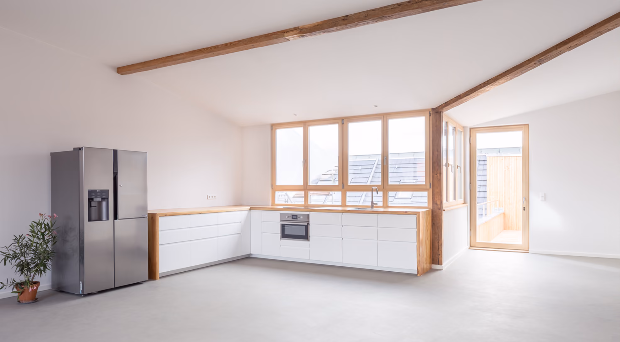 Offene Küche unter Dachschräge mit sichtbaren Holzbalken, weißer Einbauküche und raumhohen Sprossenfenstern – Projekt Dachaufstockung Berlin