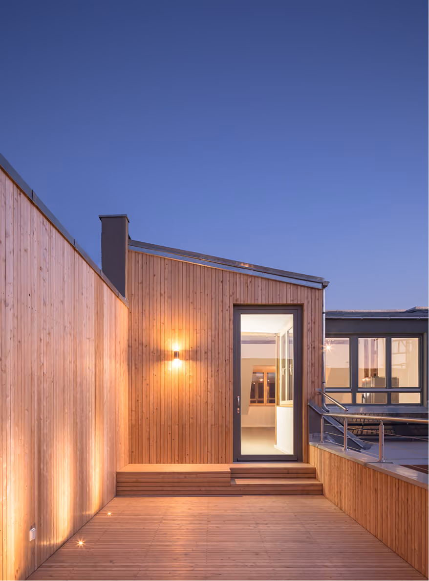 Außenansicht der Dachaufstockung mit Holzfassade, beleuchtetem Eingang und Dachterrasse in der Abenddämmerung
