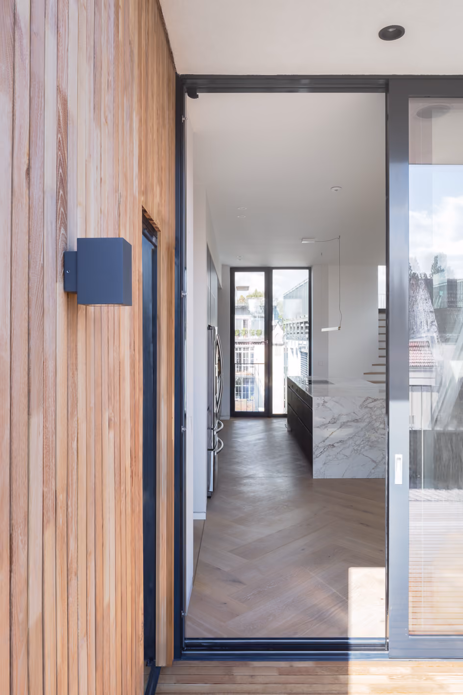 Eingangsbereich des Penthouse mit Holzlattung an der Fassade, Blick durch verglasten Flur auf Marmorwand und Dachterrasse im Hintergrund