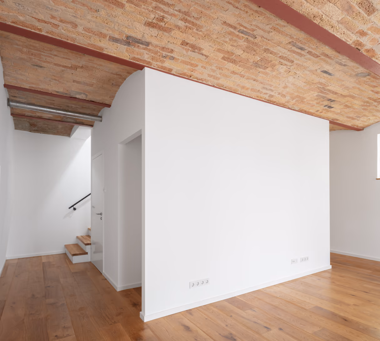 Offener Wohnraum in der Remise mit freistehender weißer Wand als Raumteiler unter sichtbarer Holzbalkendecke, Treppe im Hintergrund