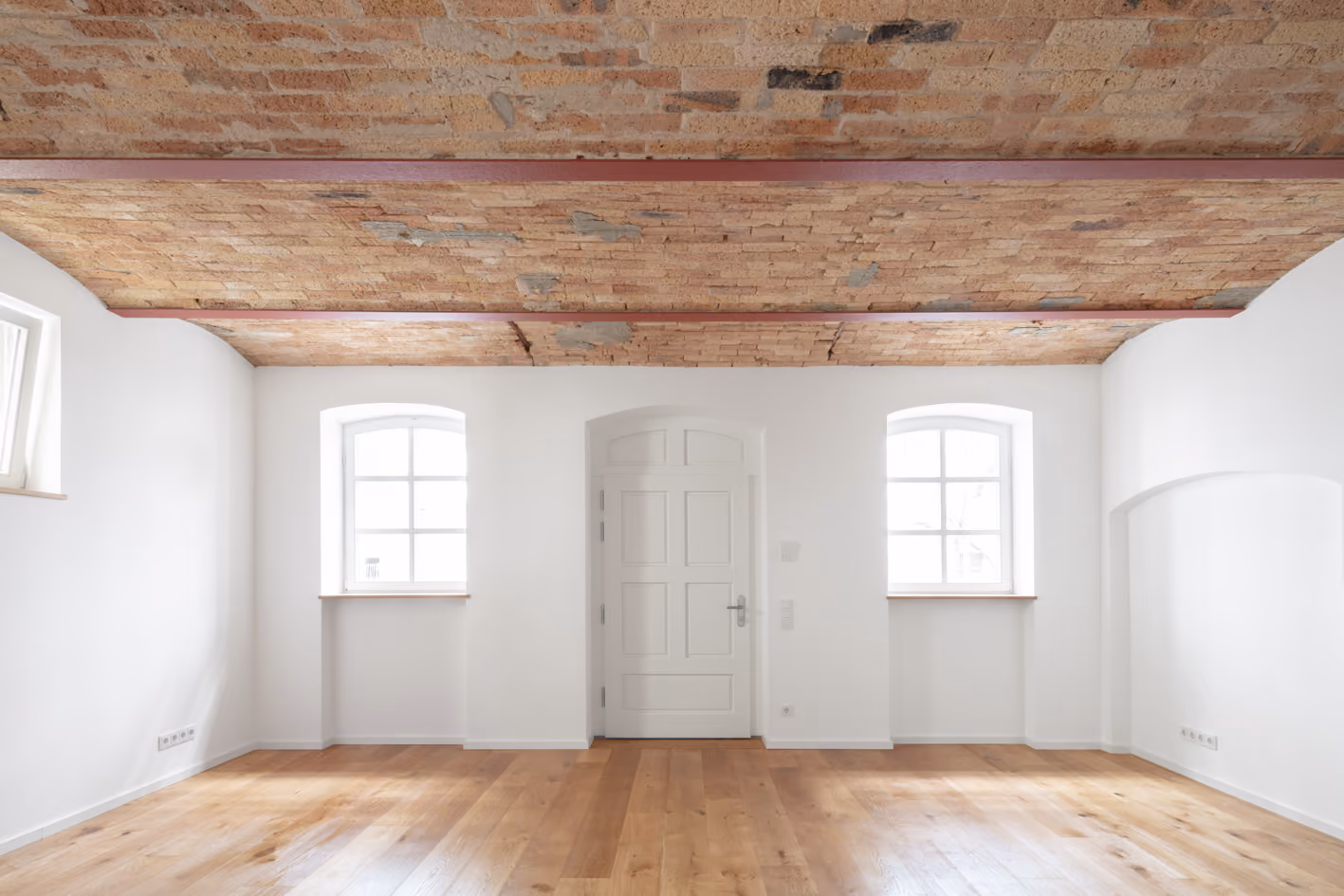 Wohnraum in der Remise mit sichtbarer historischer Holzbalkendecke und weißer Rundbogentür zwischen zwei Sprossenfenstern