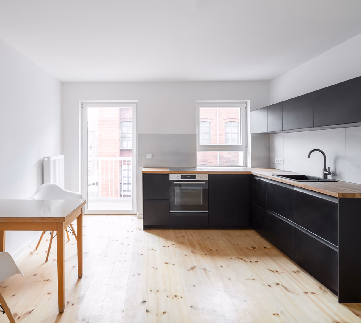 Offene Küche in der Maisonette mit schwarzer Einbauküche, hellem Holzboden und Essplatz vor Fensterfront zum Balkon