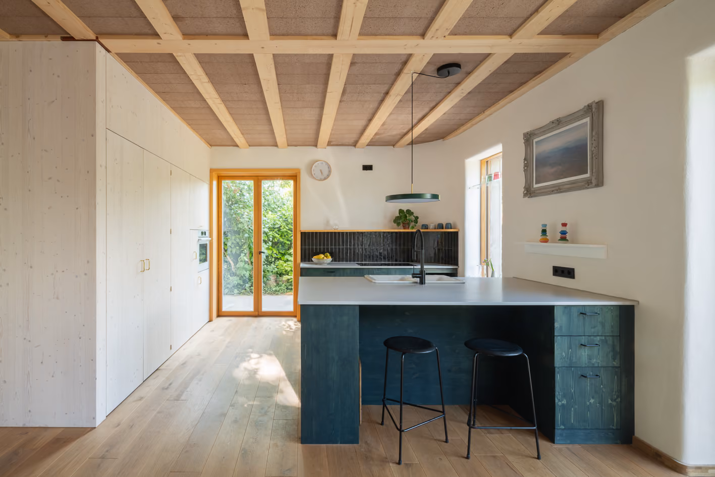 Offene Küche im Holz-Lehm-Haus mit petrolfarbener Kücheninsel, sichtbarer Holzbalkendecke und Terrassentür zum Garten