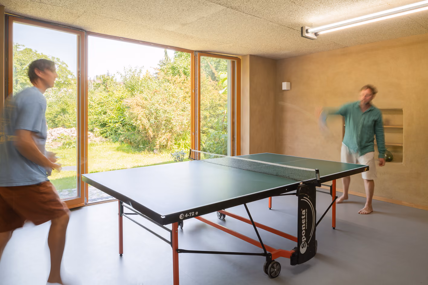 Tischtennisraum im Nebengebäude des Holz-Lehm-Hauses mit großer Glasfront zum Garten und Lehmwänden