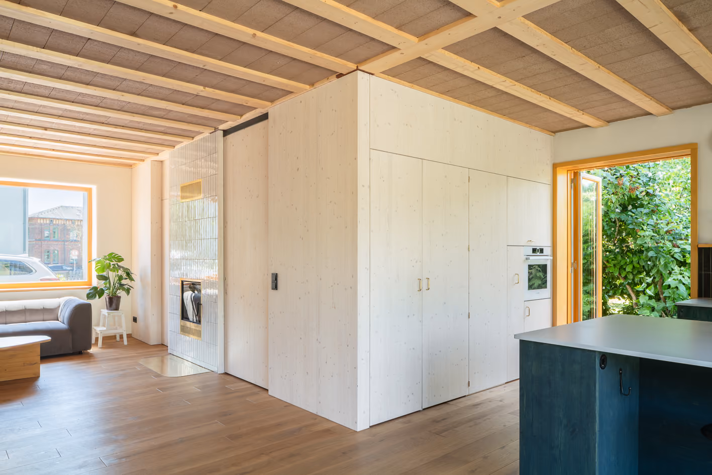 Offener Wohnbereich im Holz-Lehm-Haus mit weißem Einbaukubus, sichtbarer Holzbalkendecke und grüner Küchenzeile im Hintergrund