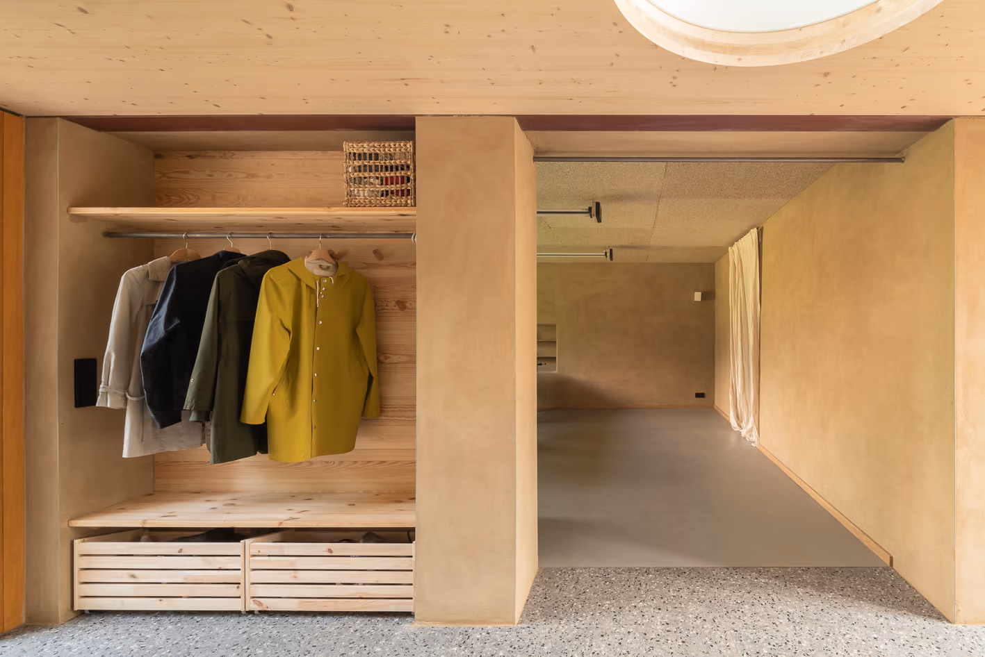 Eingebaute Garderobe aus hellem Holz im Holz-Lehm-Haus mit offener Kleiderstange und Durchgang zum Nebenraum mit Oberlicht