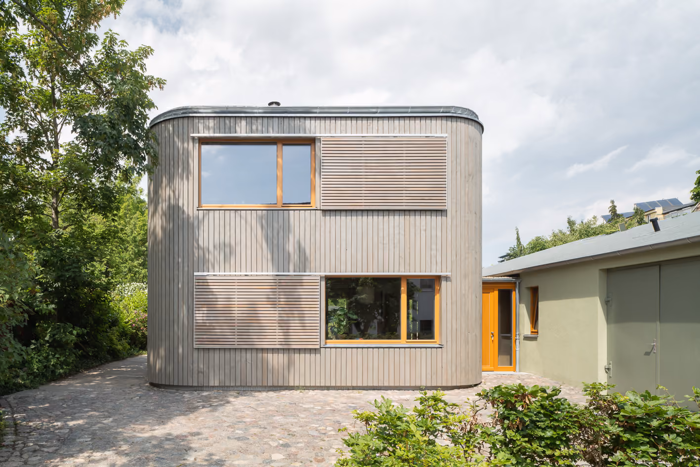Frontale Außenansicht des Holz-Lehm-Hauses mit geschwungener Holzfassade und abgerundeten Ecken im Grünen