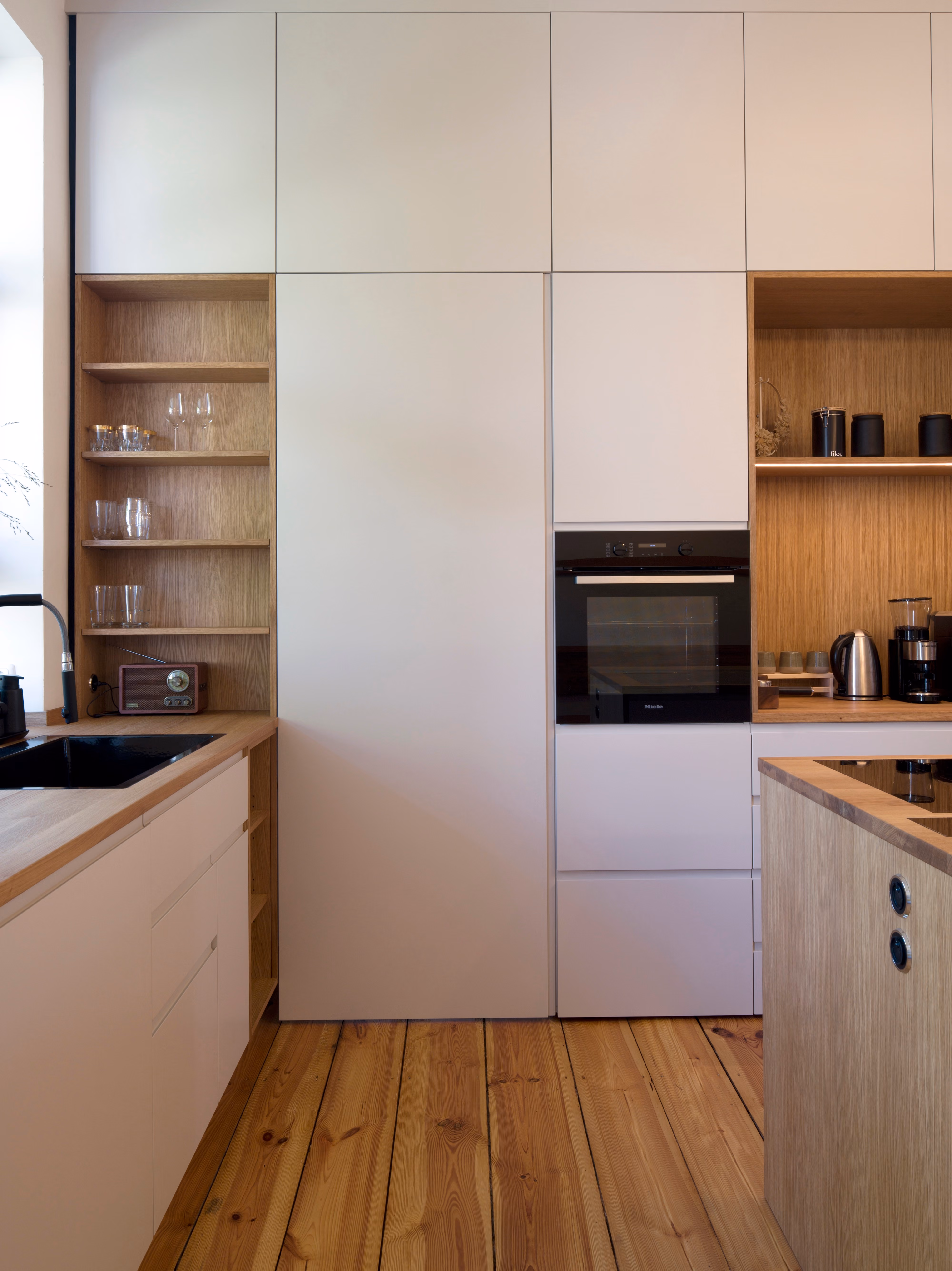 Einbauküche in der Altbauwohnung Schwerin mit weißen Fronten, integriertem Backofen und offenem Holzregal als Kontrast