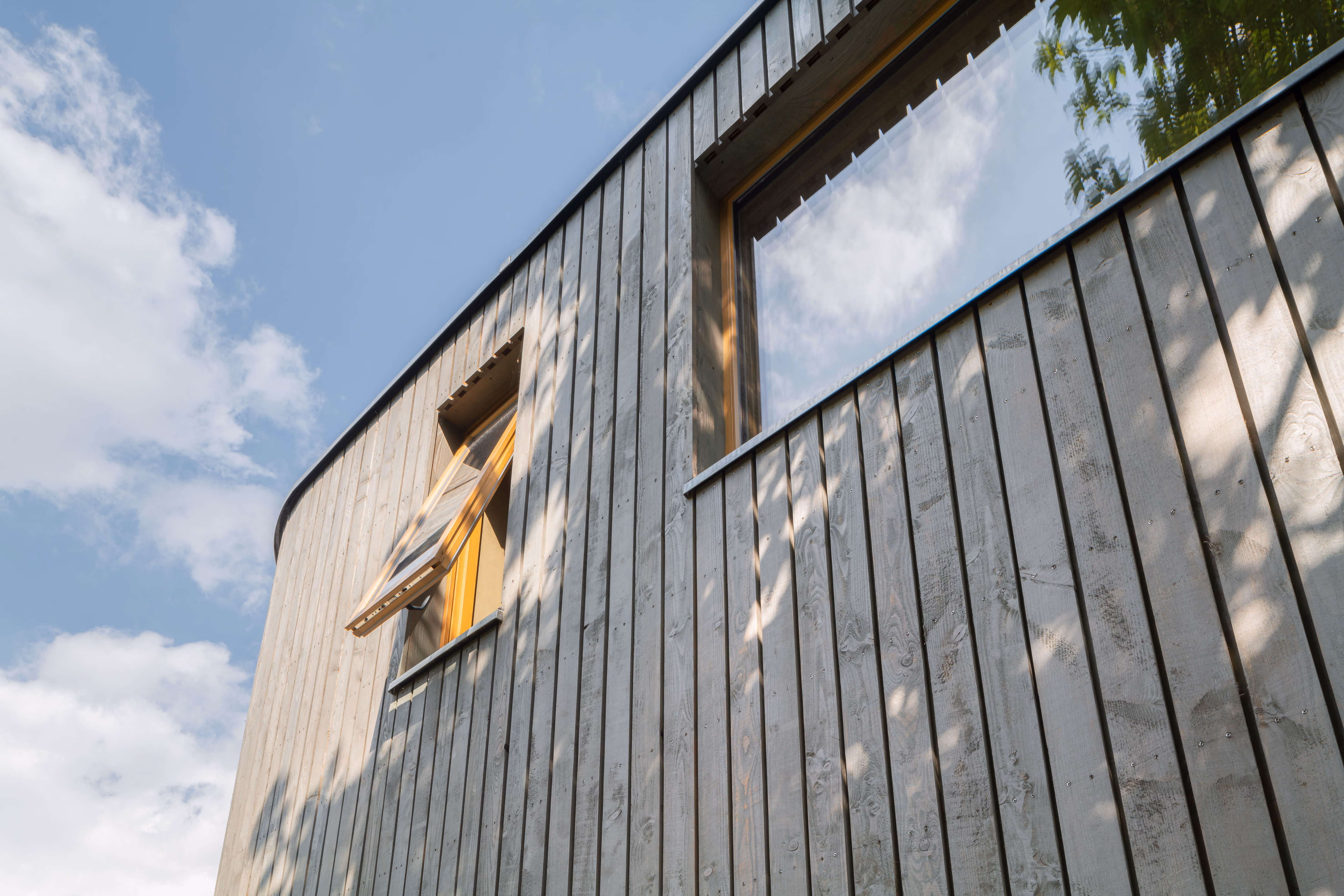 Untersicht der geschwungenen Holzfassade des Holz-Lehm-Stroh-Hauses mit vertikaler Schalung aus vergrautem Holz vor blauem Himmel
