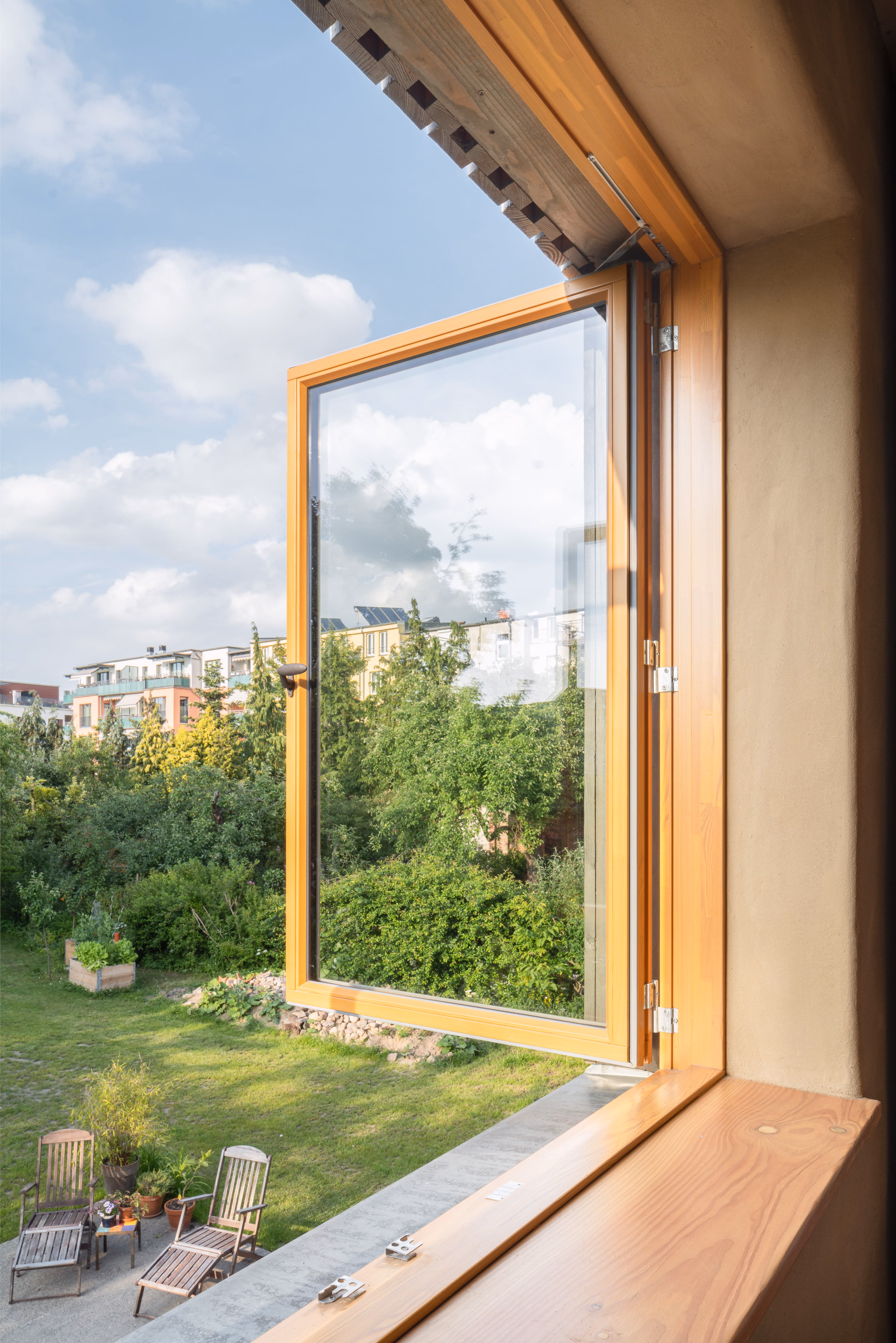 Geöffnetes Holzfenster mit Lehmputz-Laibung im Holz-Lehm-Stroh-Haus, Blick über einen grünen Garten auf die Nachbarbebauung