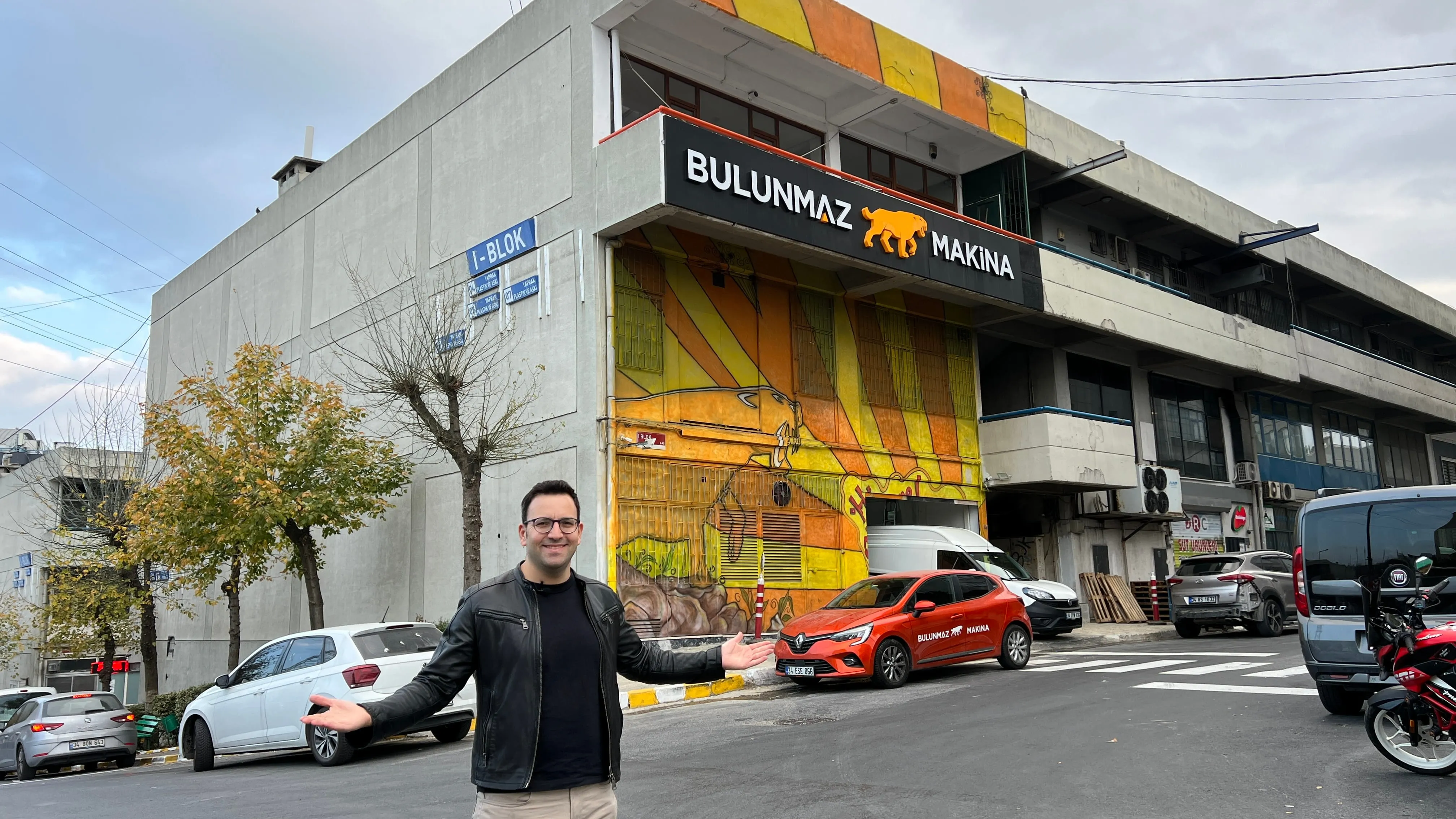 Man in black jacket gestures outside Bulunmaz Makina building with yellow mural and branded car.