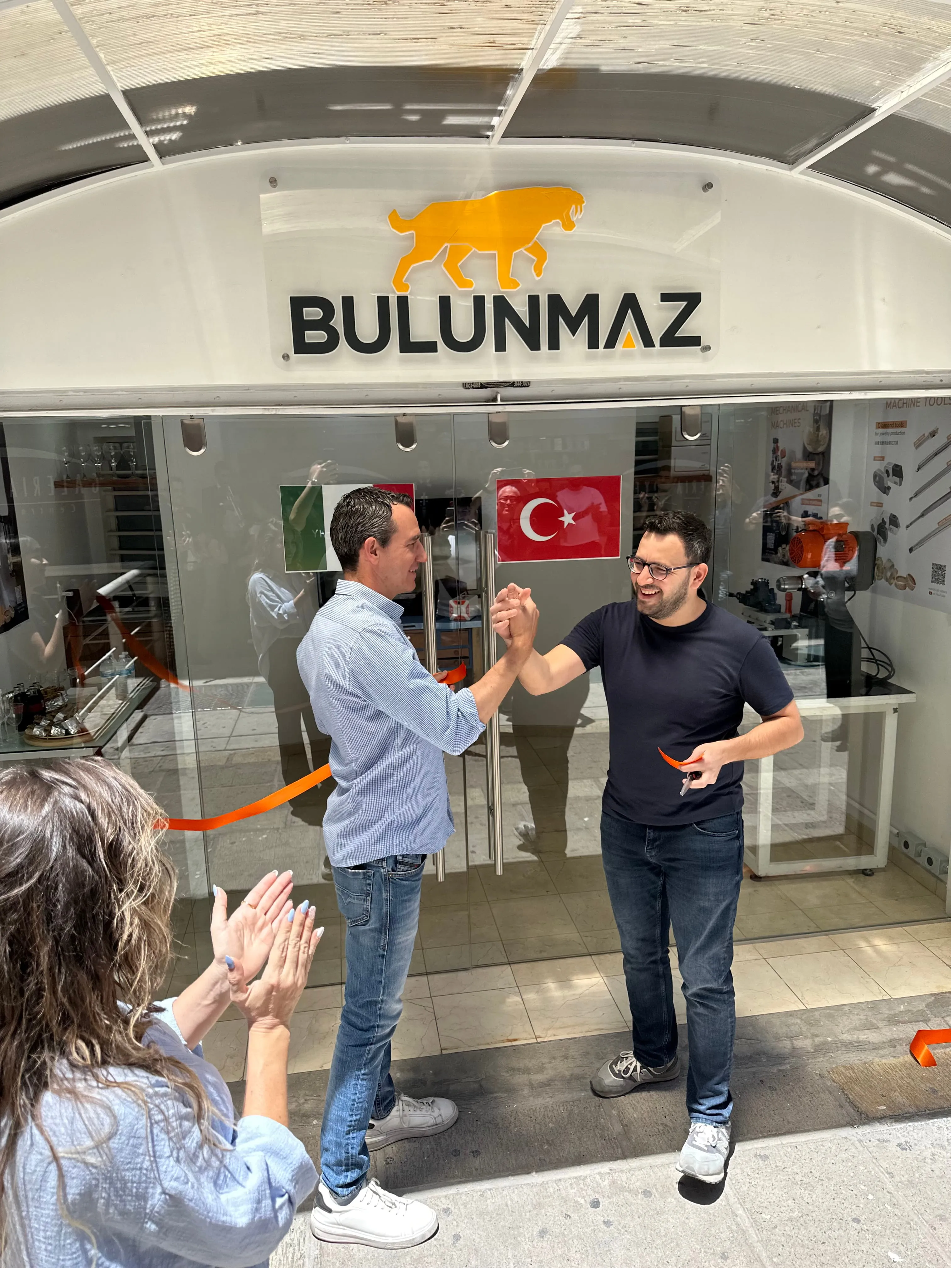 Two men shaking hands in front of glass doors with Bullunmaz sign and flags, a woman clapping nearby.
