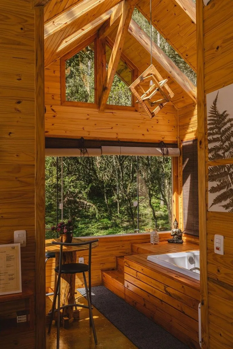 Cozy wooden cabin interior with large windows, a bathtub, seating area, and decor including a Buddha statue and flowers. Natural light floods in.
