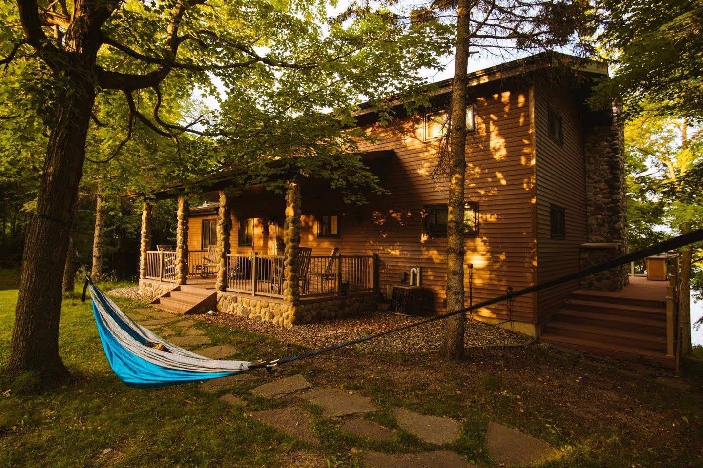 A cozy wooden cabin surrounded by trees, featuring a hammock, stonework, and a porch bathed in warm sunlight. Perfect for relaxation.
