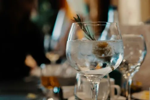 Close-up of a glass of clear liquid with a sprig of herbs, surrounded by blurred glasses on a table, creating a cozy dining atmosphere.