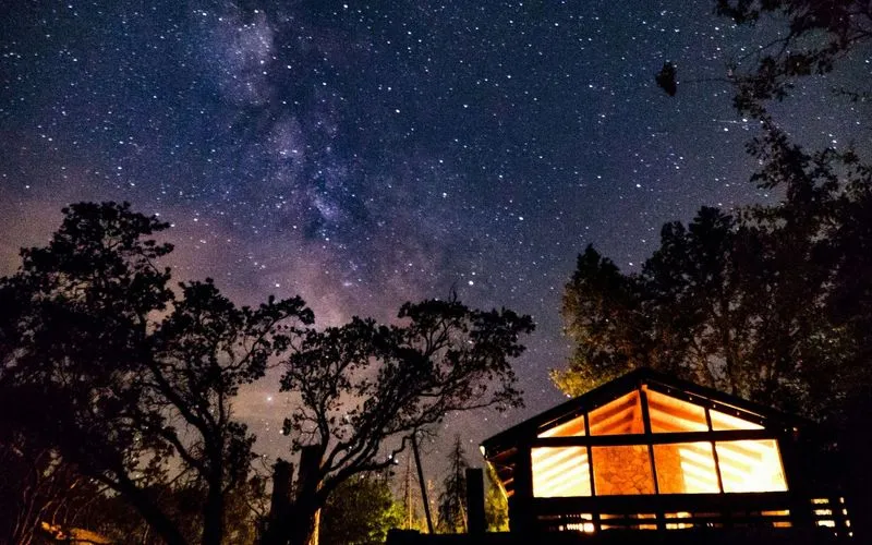 Starry night sky with Milky Way and a lit cabin surrounded by trees.