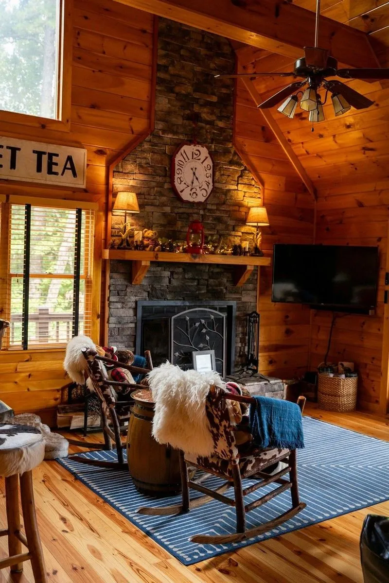 Cozy wooden cabin room with stone fireplace, rocking chairs, striped rug, and ceiling fan.