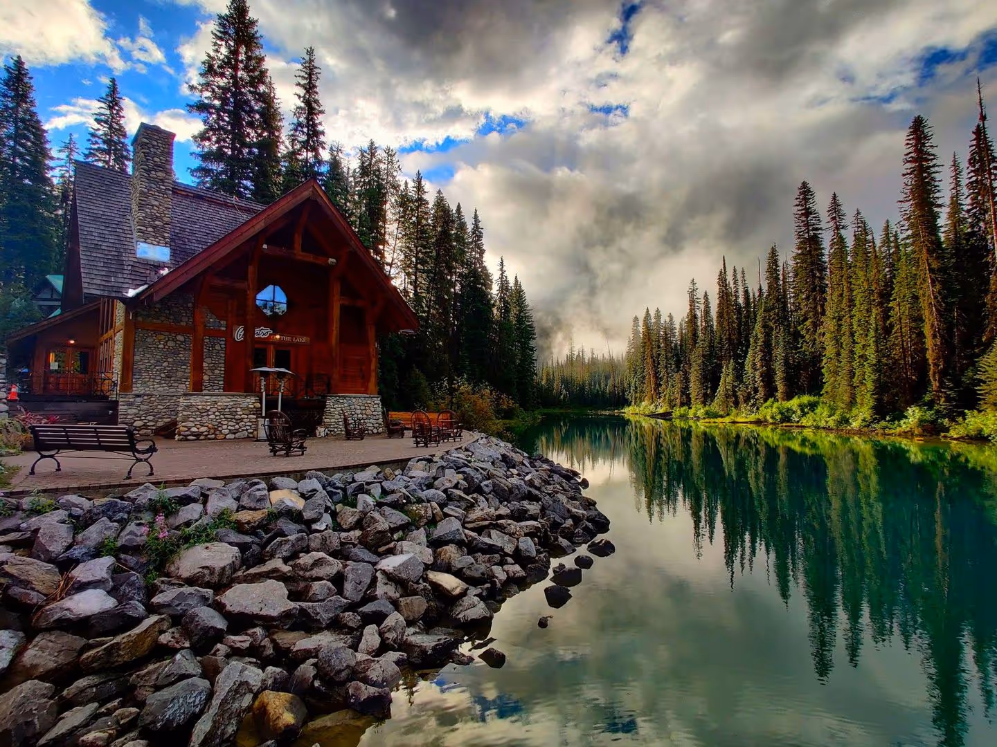 Stone and wood cabin by a rocky lake shore with tall pine trees reflecting in calm water.
