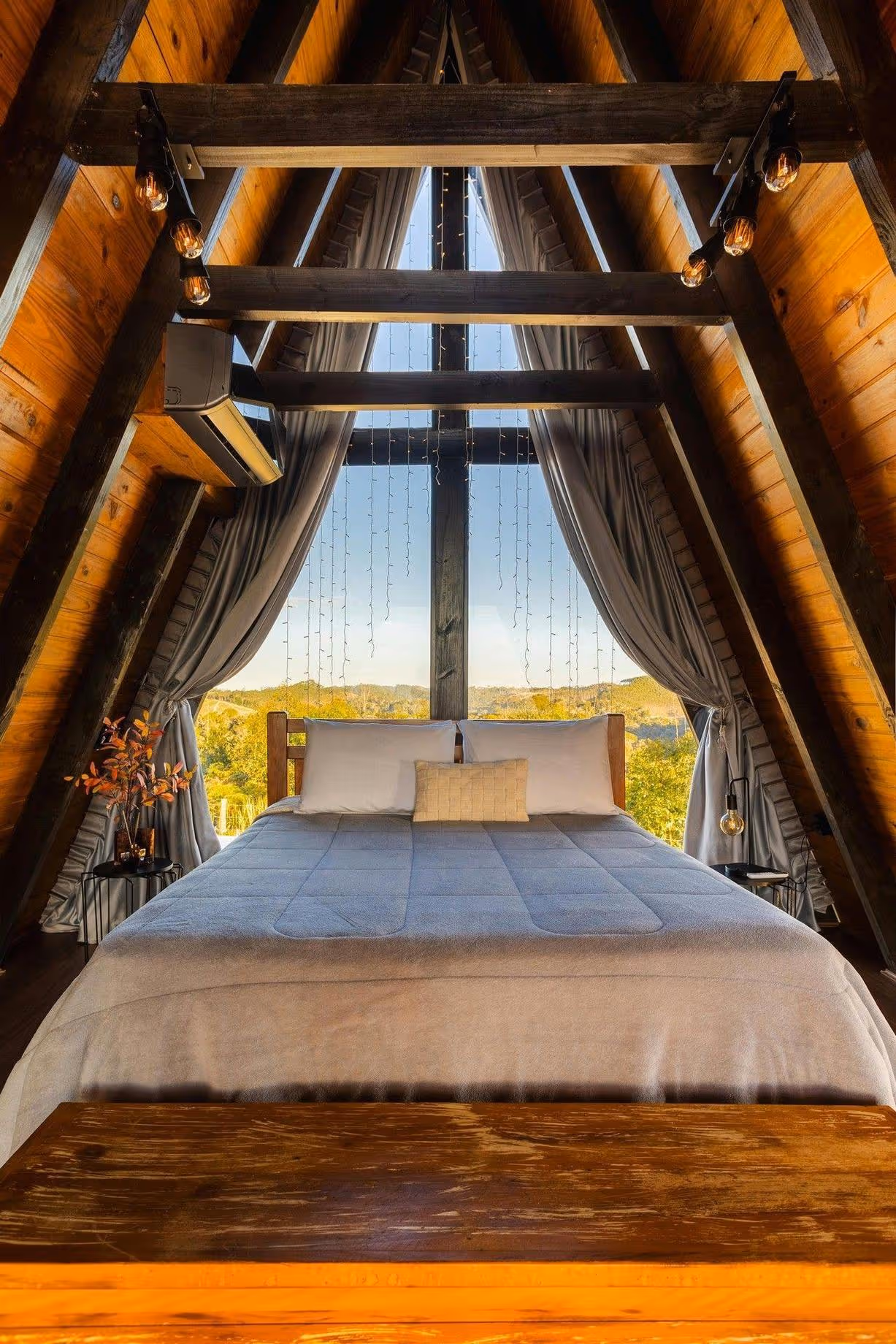 A-frame cabin bedroom with a large bed facing a window showing trees and hills outside.