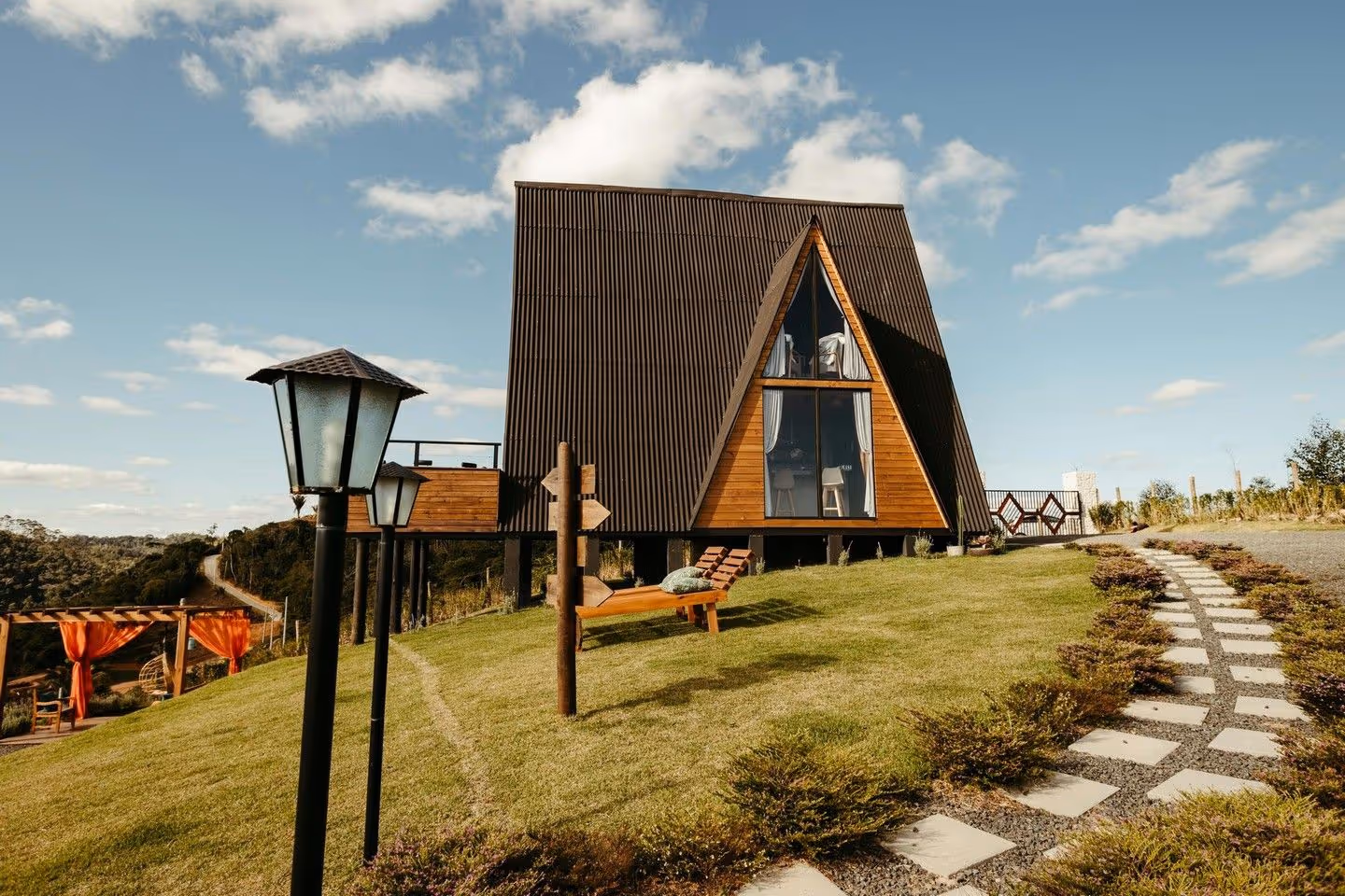 A-frame cabin with large windows on a grassy hill with a pathway and lampposts under a blue sky.