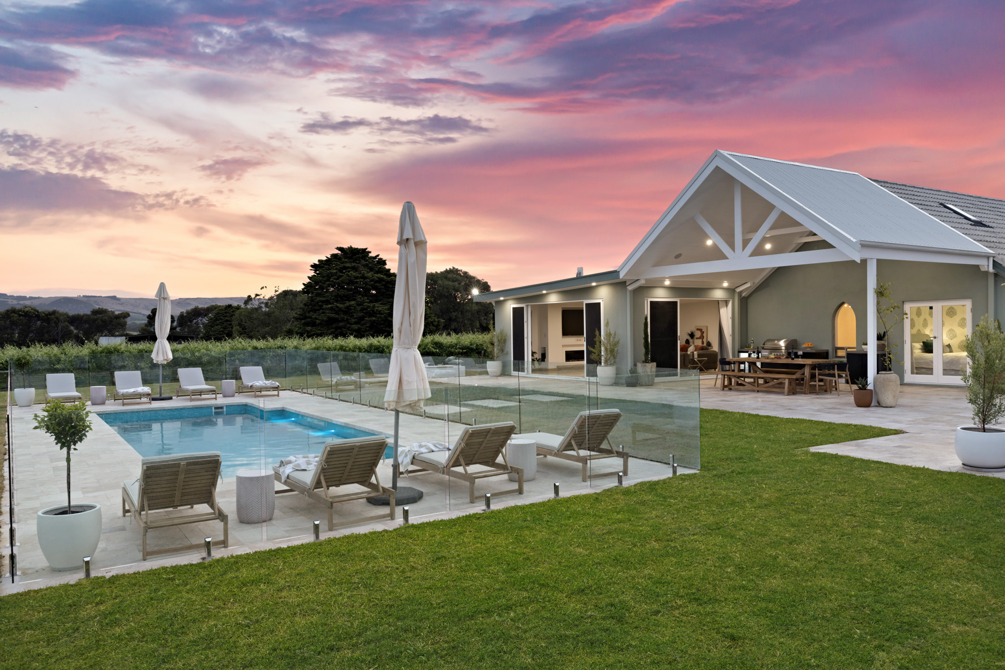 Modern house with glass-fenced pool, lounge chairs, and vibrant sunset sky.