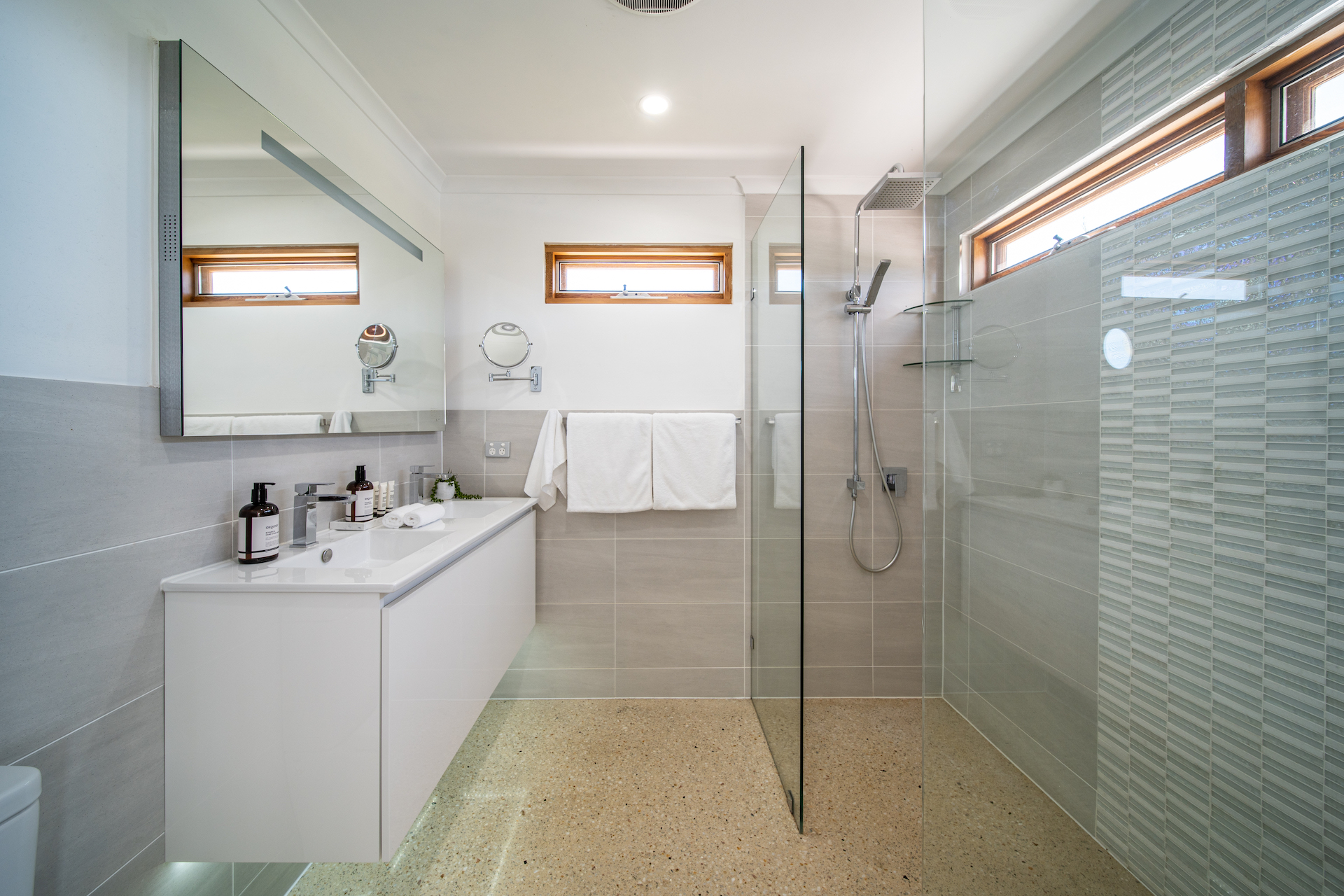 Modern bathroom with floating vanity, large mirror, glass shower, and two windows with wooden frames.