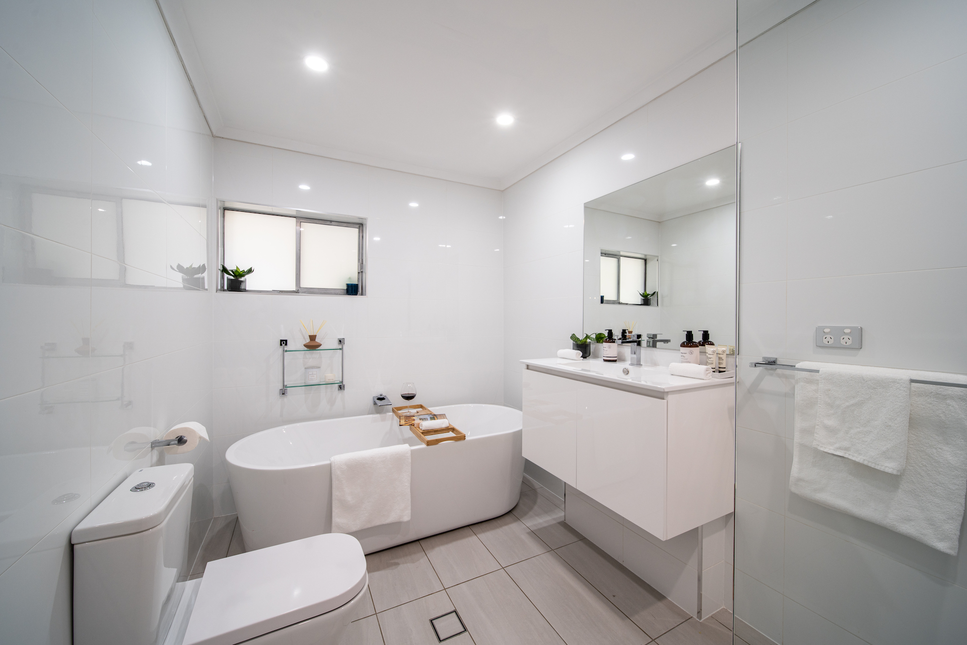 Modern white bathroom with bathtub, toilet, large mirror, floating vanity, and towel racks.