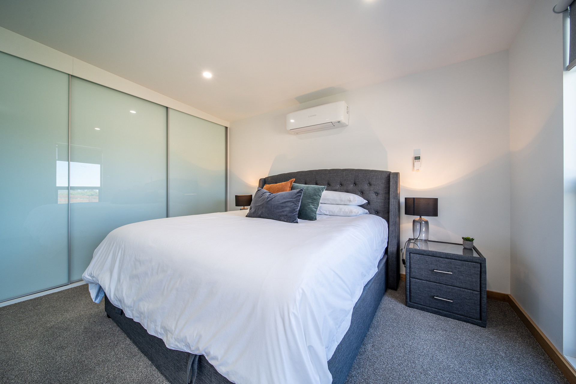 Modern bedroom with gray upholstered bed, white bedding, colorful pillows, nightstand, and lamps.