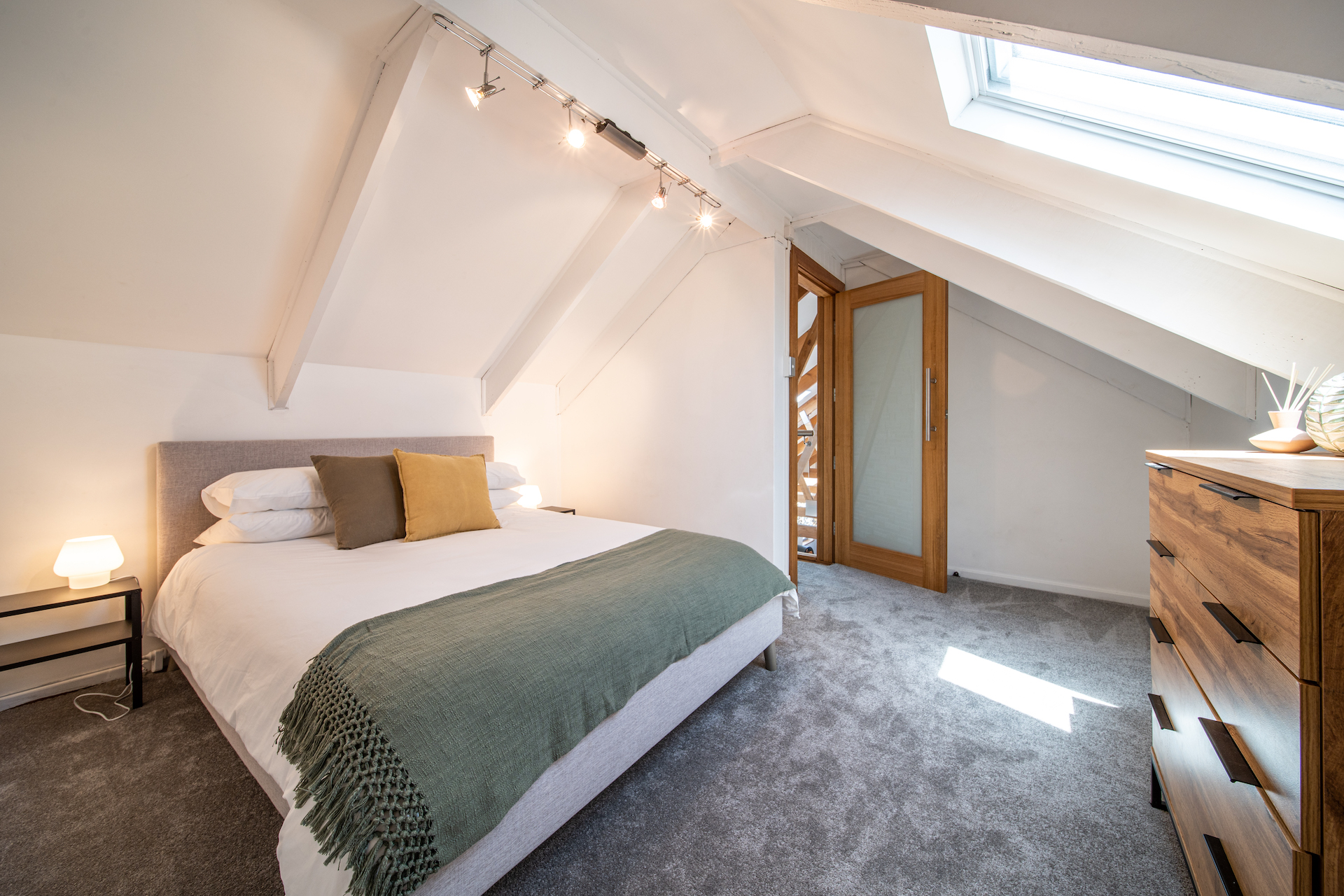 Bright attic bedroom with a bed, green throw blanket, wooden dresser, and skylight window.