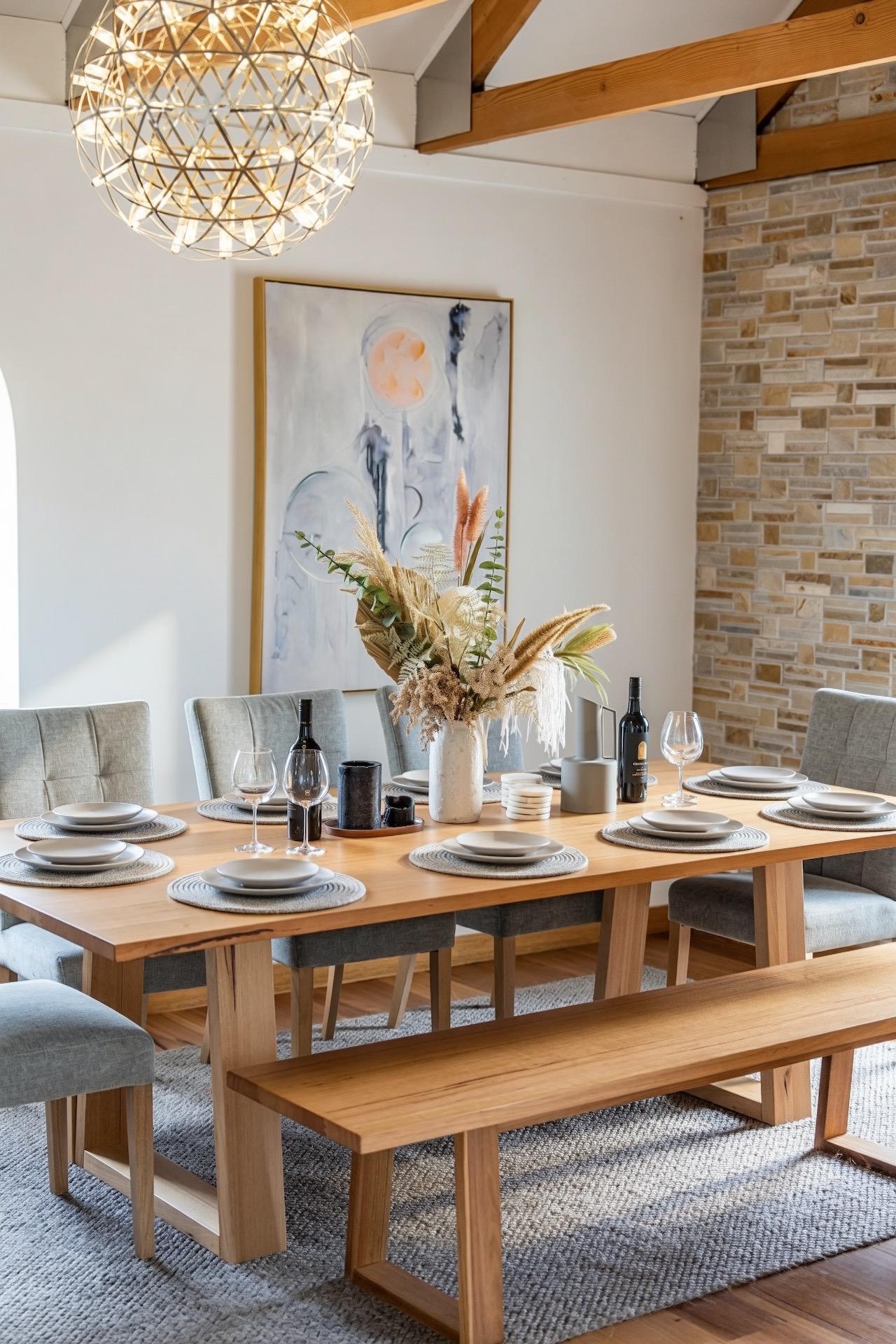 Dining table set with plates, wine glasses, two bottles, and a vase of dried flowers in bright room.