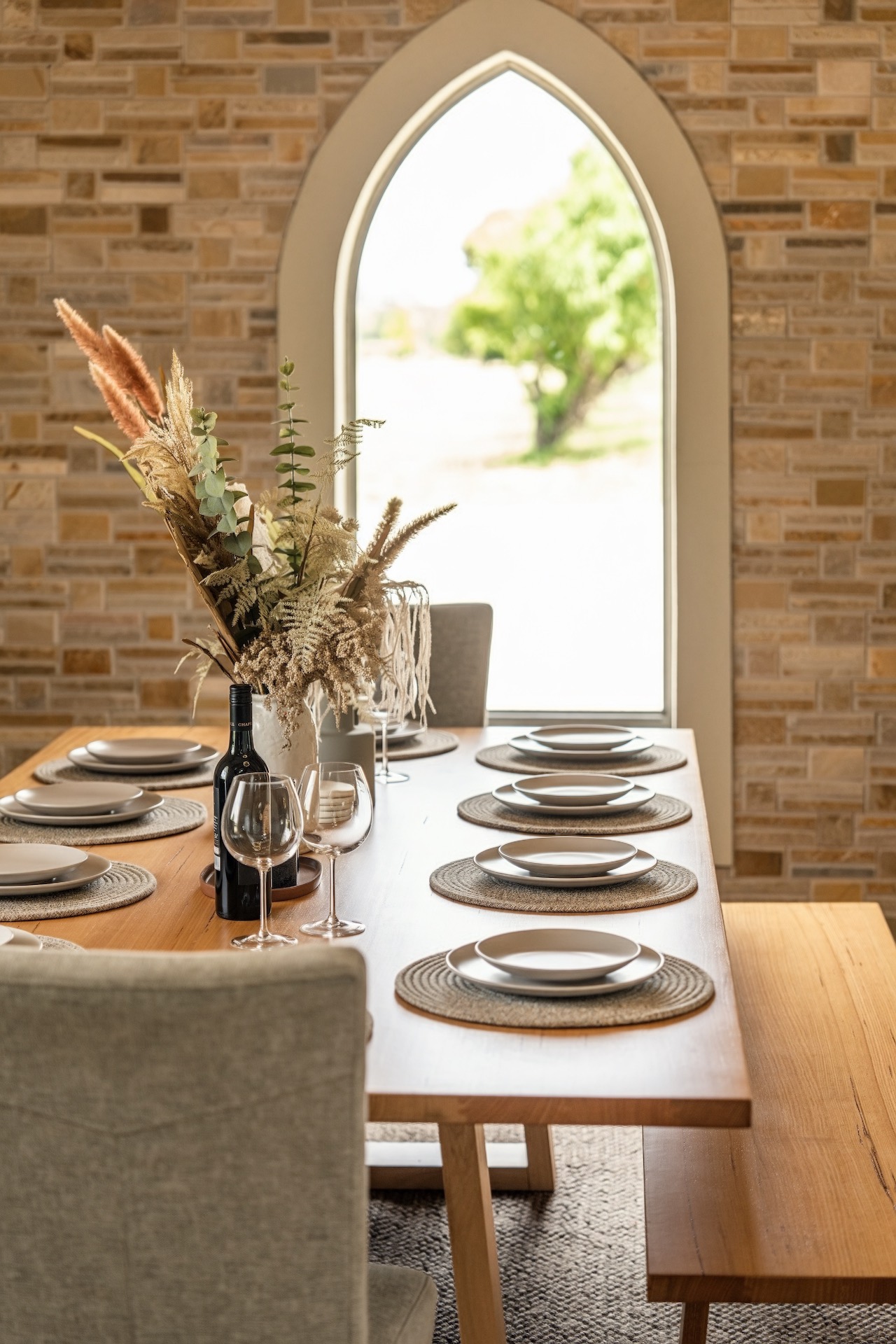 Wooden dining table set with neutral plates and woven mats near an arched window and brick wall.