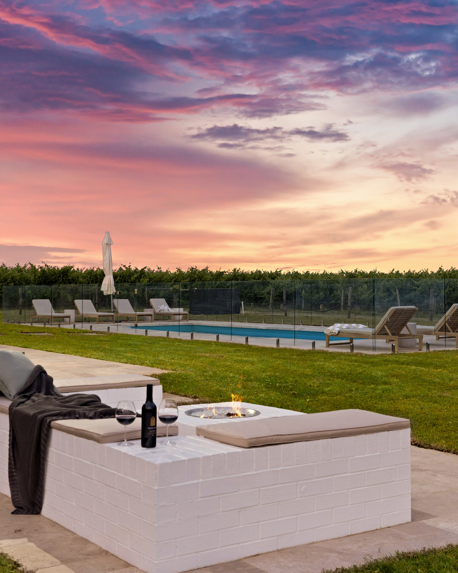 Outdoor seating with cushioned benches, fire pit, wine glasses, and a view of pool and sunset sky.