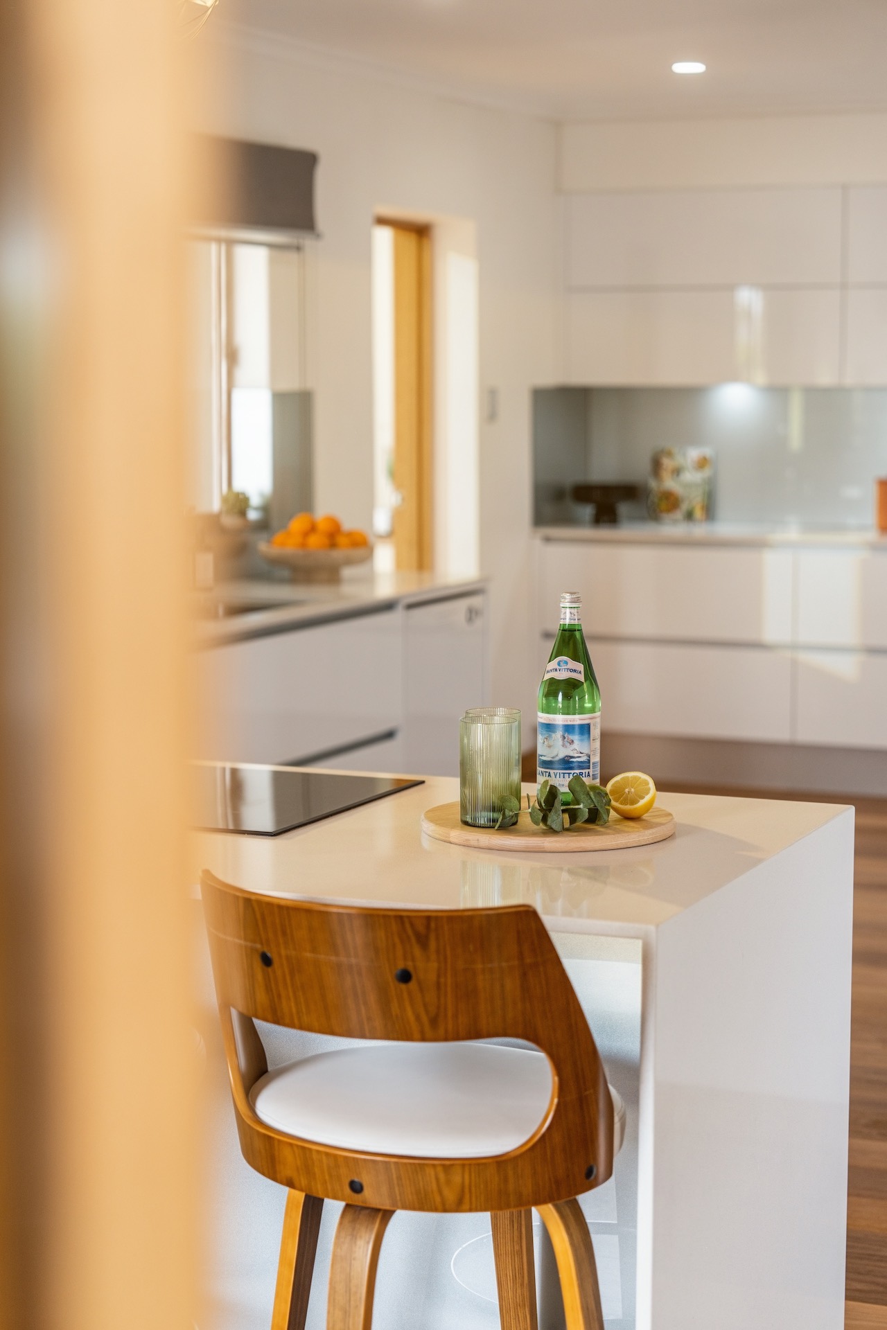 Wooden bar stool in a modern kitchen with a bottle, glass, lemon, and leaves on the counter.
