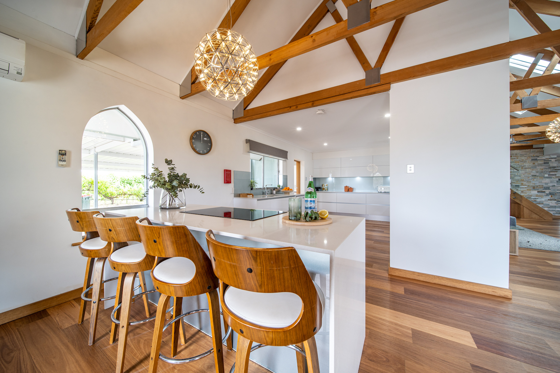 Modern white kitchen with wooden bar stools, wooden ceiling beams, and pendant light fixture.