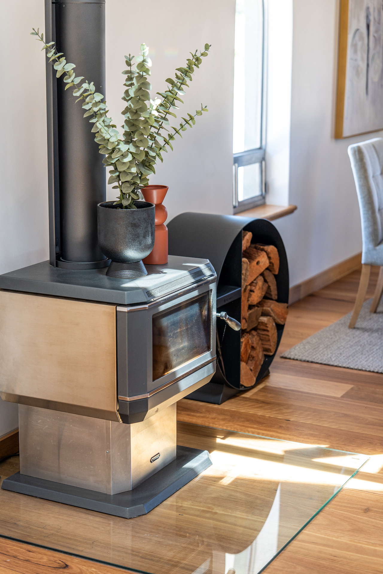 Modern metal wood stove with plants on top and firewood stacked beside it on a wooden floor.