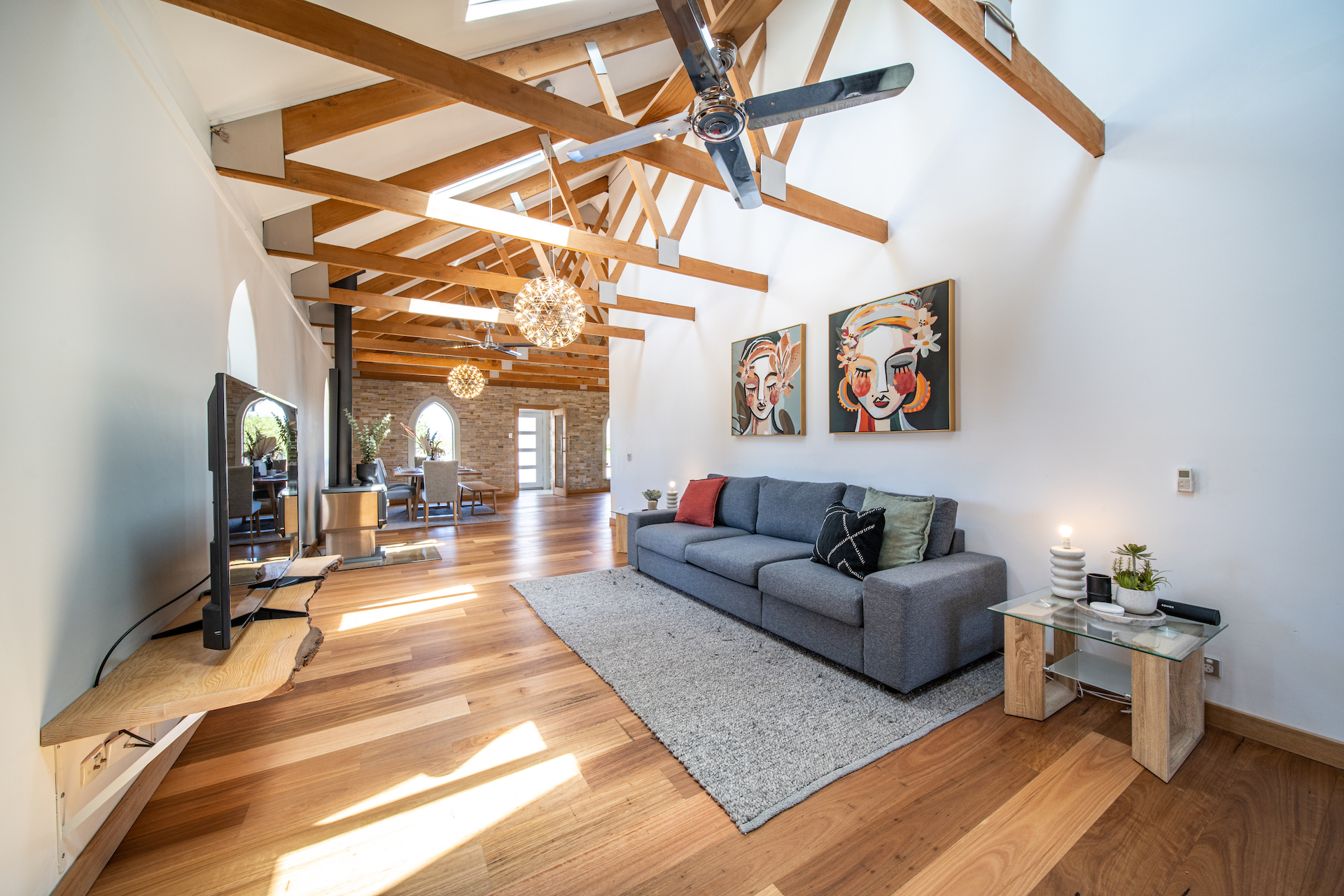 Modern living room with wooden floor, gray sofa, colorful art, exposed wooden beams, and ceiling fans.