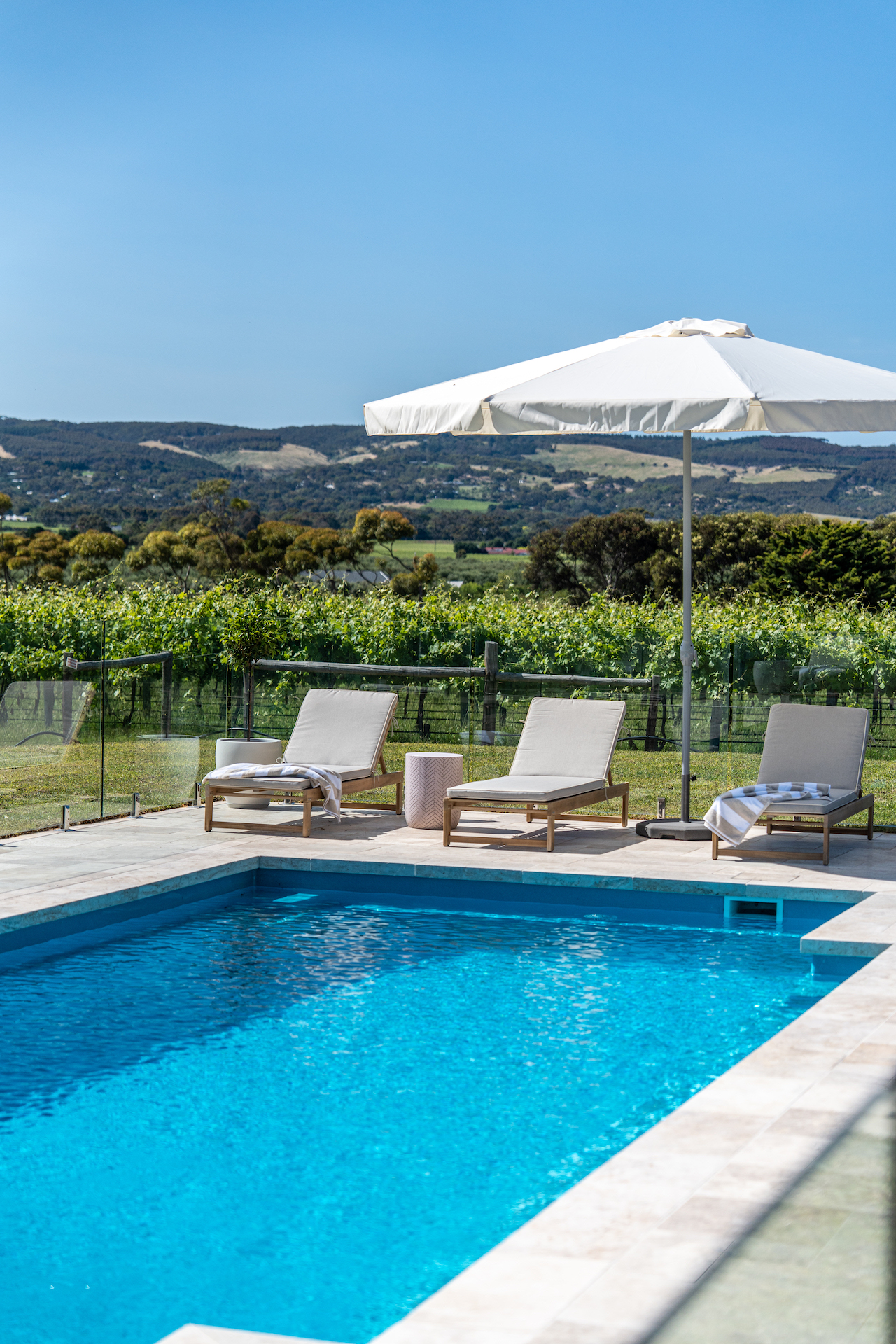 Outdoor pool with lounge chairs, towels, a white umbrella, glass fence, and vineyard hills in background.