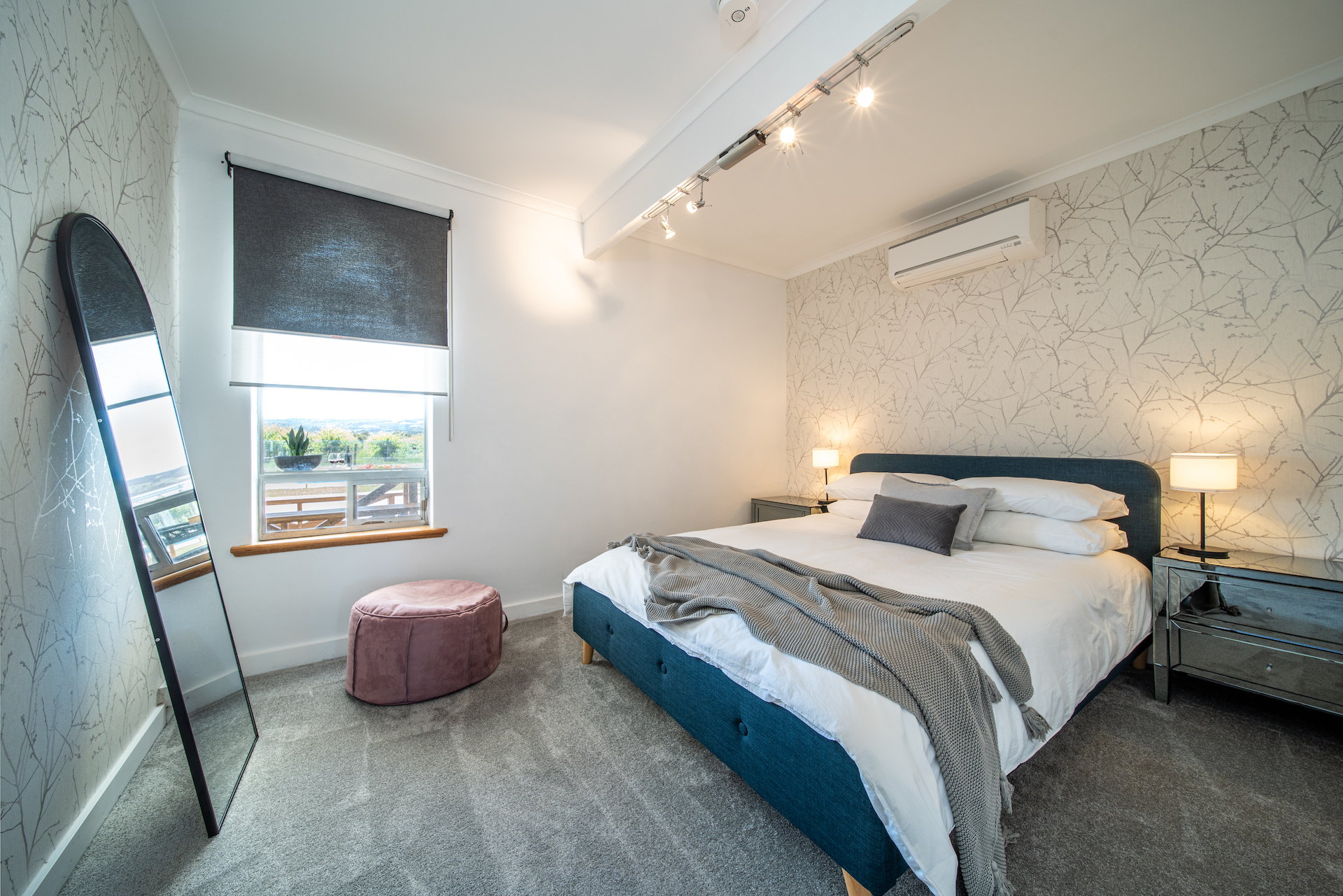 Modern bedroom with queen bed, bedside lamps, full-length mirror, and window with grey blinds.