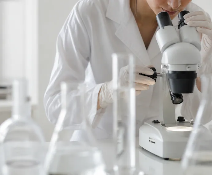 Scientist in white coat and gloves looking into a microscope in a lab with glassware.