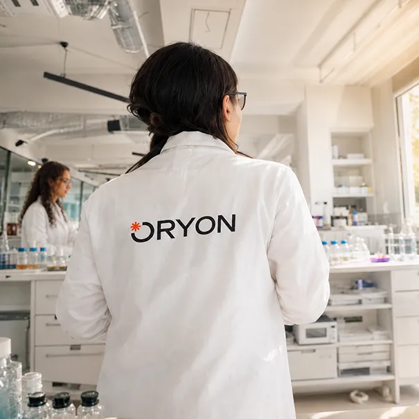Scientist in white ORYON lab coat in a bright laboratory with glassware and another person in background.