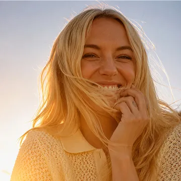 Smiling blonde woman with long hair holding strands near her face in warm sunlight.